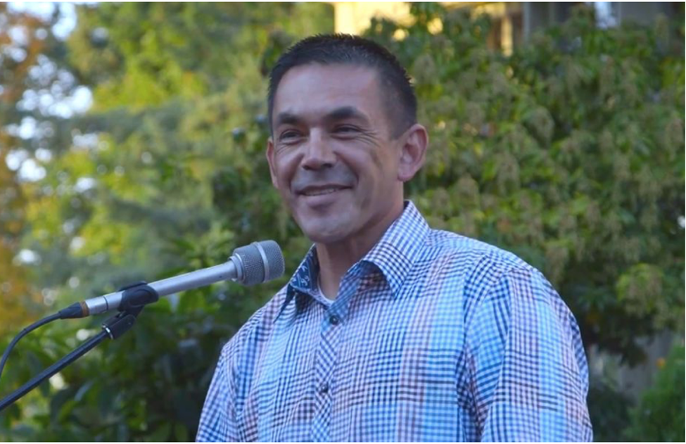 Jay Julius is smiling behind a podium microphone. He is wearing a plaid shirt. There are trees in the background.