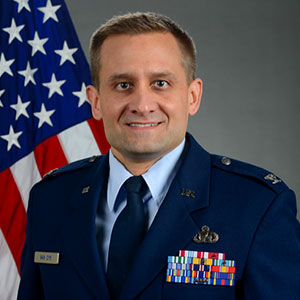 Lieutenant Colonel Greg Van Dyk is a white male with short sandy brown hair. he wears a dark blue US military uniform. The US flag is in the background.