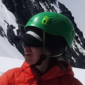 Deborah staring off into the distance wearing a green climbing helmet, and an orange coat standing on the mountaintop