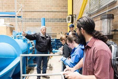 students looking at instructor standing in front of blue pipe