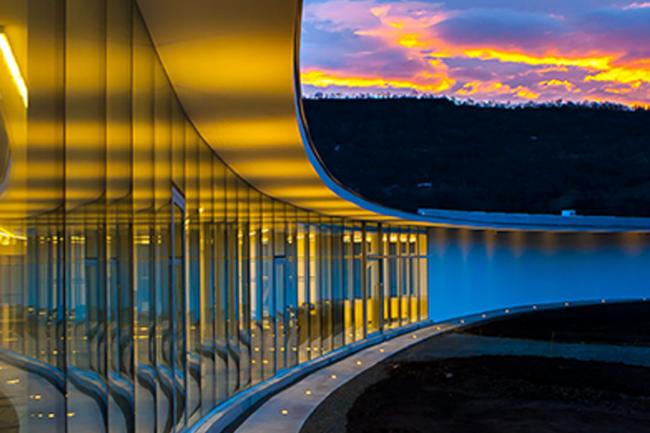 Clouds and the golden light of dusk reflect dramatically on the curved glass surfaces of a modern building.